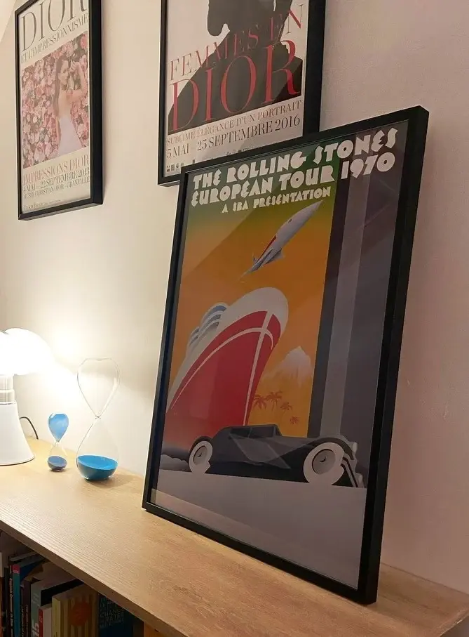 Cadre Eva noir posé contre un mur, affiche de concert vintage, lampe et sabliers